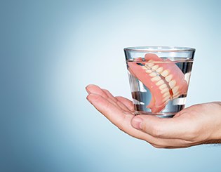 A hand holding a glass of clear liquid with dentures in front of a faded blue background